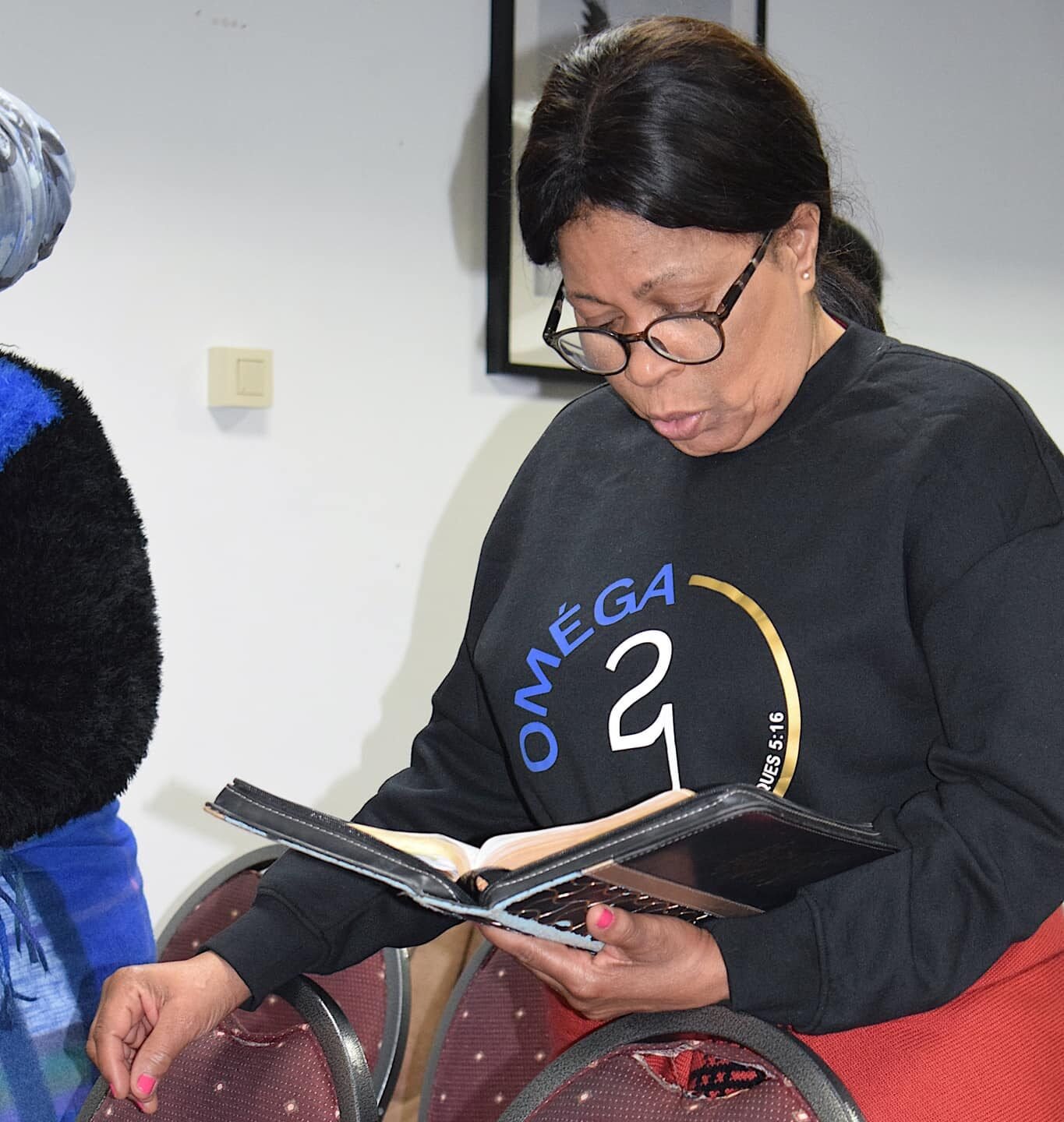 A woman in a black sweater with the 'OMEGA 21' logo reading a book intently in a church setting