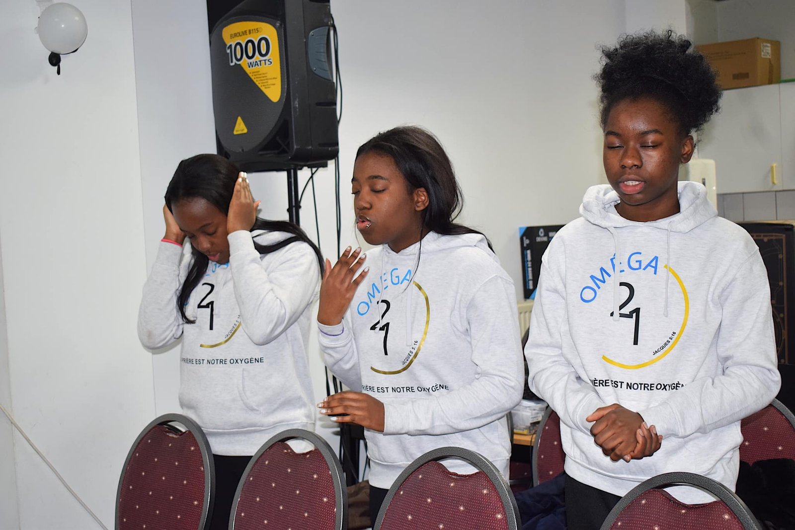 Young congregation members in prayer, wearing 'Omega 21' hoodies at Église La Bonne Semence