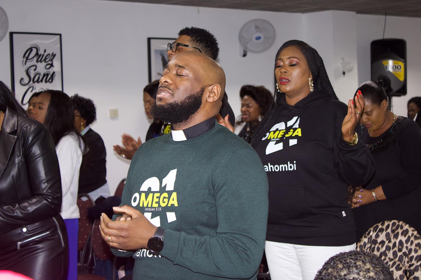 Members of Église La Bonne Semence in prayer, wearing 'Omega' hoodies during a church service.