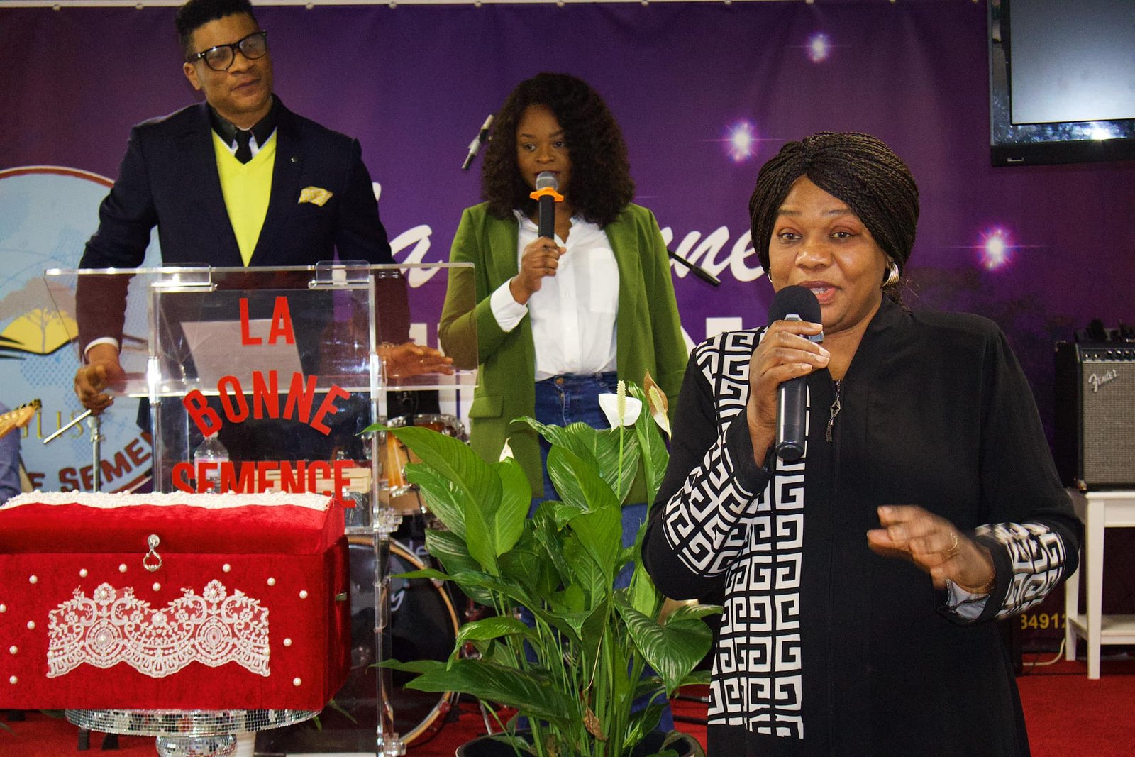 Senior woman speaking passionately at a podium, with other church leaders in the background at Église La Bonne Semence