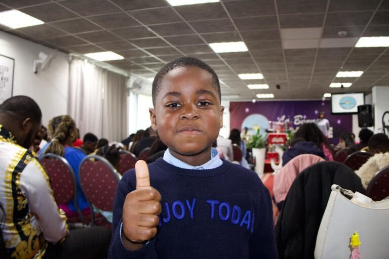 Young boy giving a thumbs up in a church setting, wearing a sweater with the words 'JOY TODAY