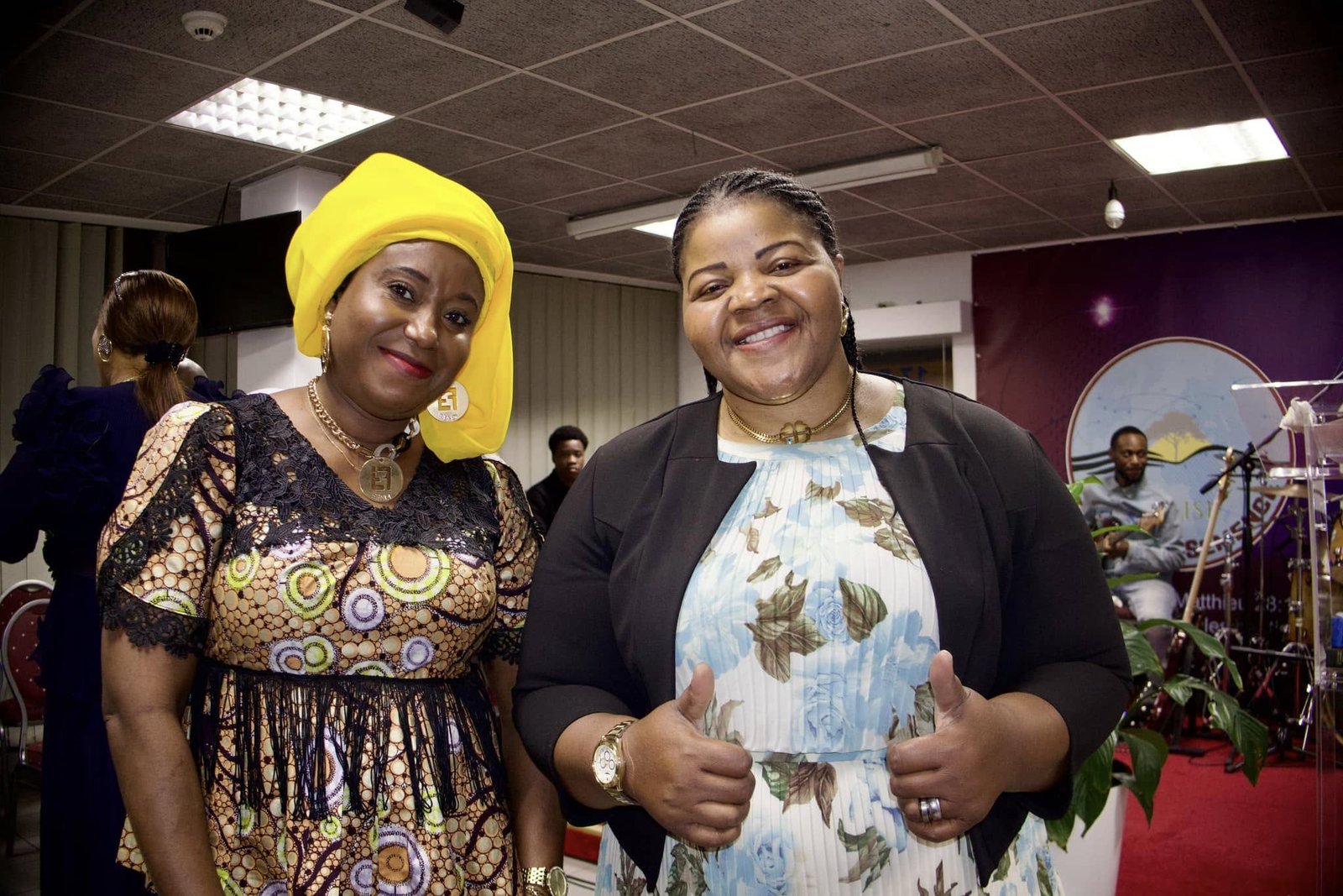 Two women smiling at a church event, one wearing a yellow headscarf and a patterned dress, the other in a floral dress.