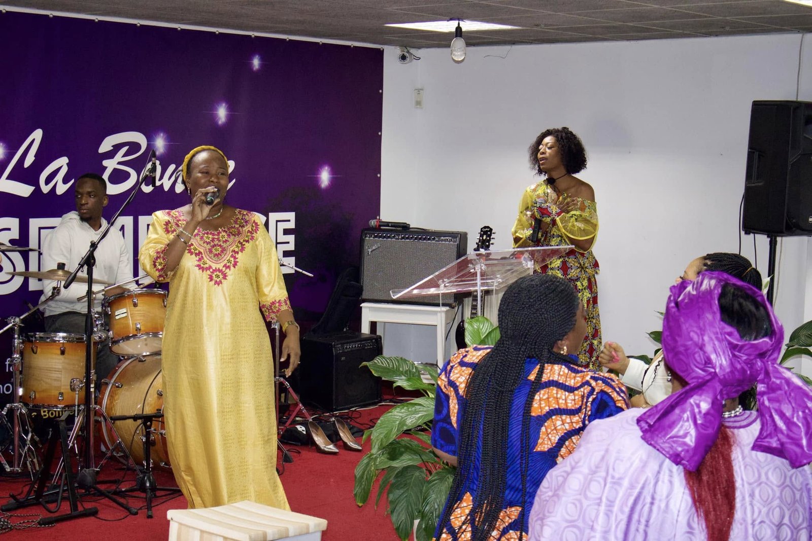 Two women singing at a church service at Église La Bonne Semence, one in a yellow dress and the other in a colorful floral dress.
