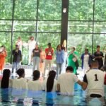 Church members singing and clapping on stage during a baptism event, with attendees watching from a water-filled baptismal pool.