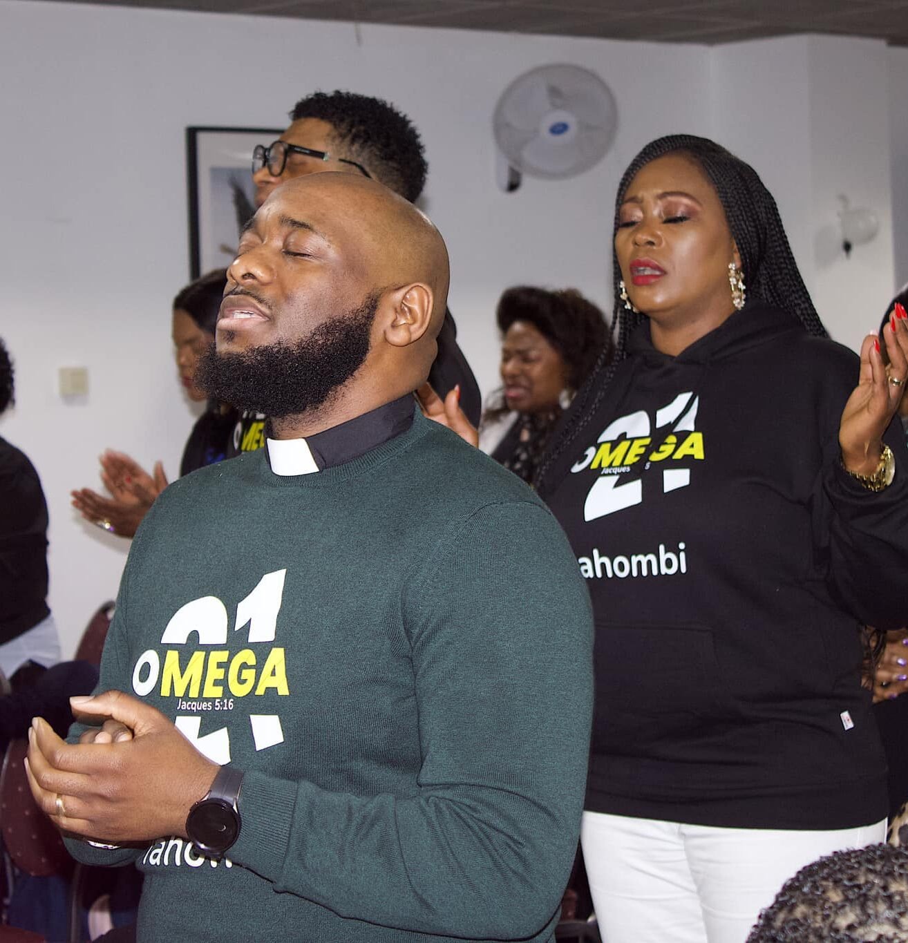 A group of adults praying and worshiping in a church, with a man wearing a green sweater and black clerical collar in the foreground