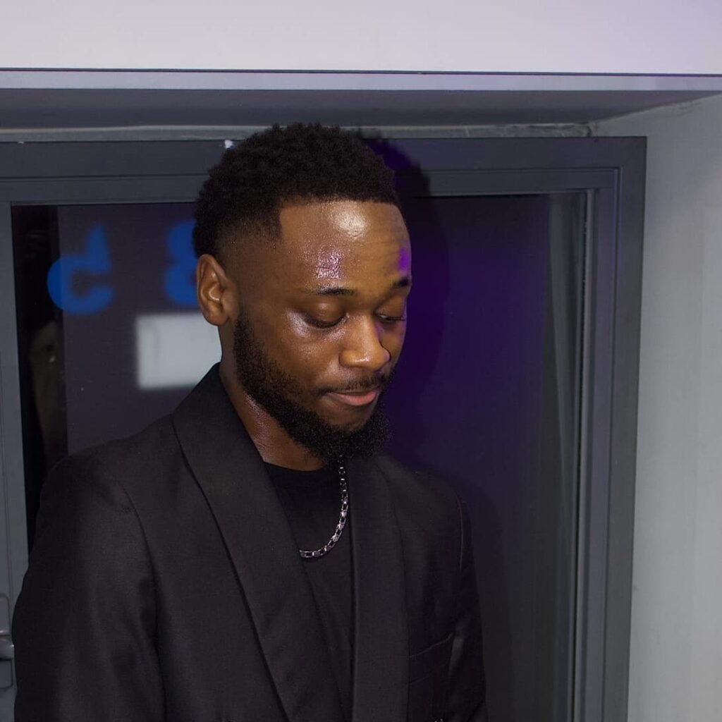 A young man, Moise Mukendi, holding a guitar, dressed in a black shirt and strap.