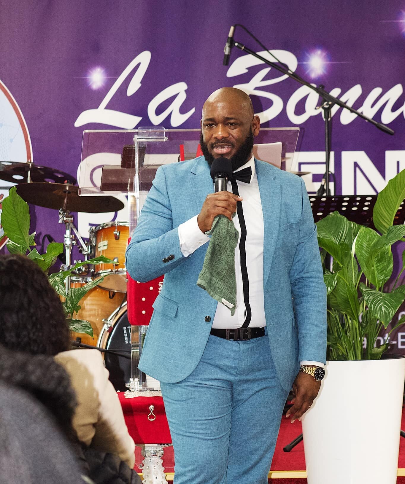 A confident man speaking at a church podium in a stylish blue suit and bow tie.