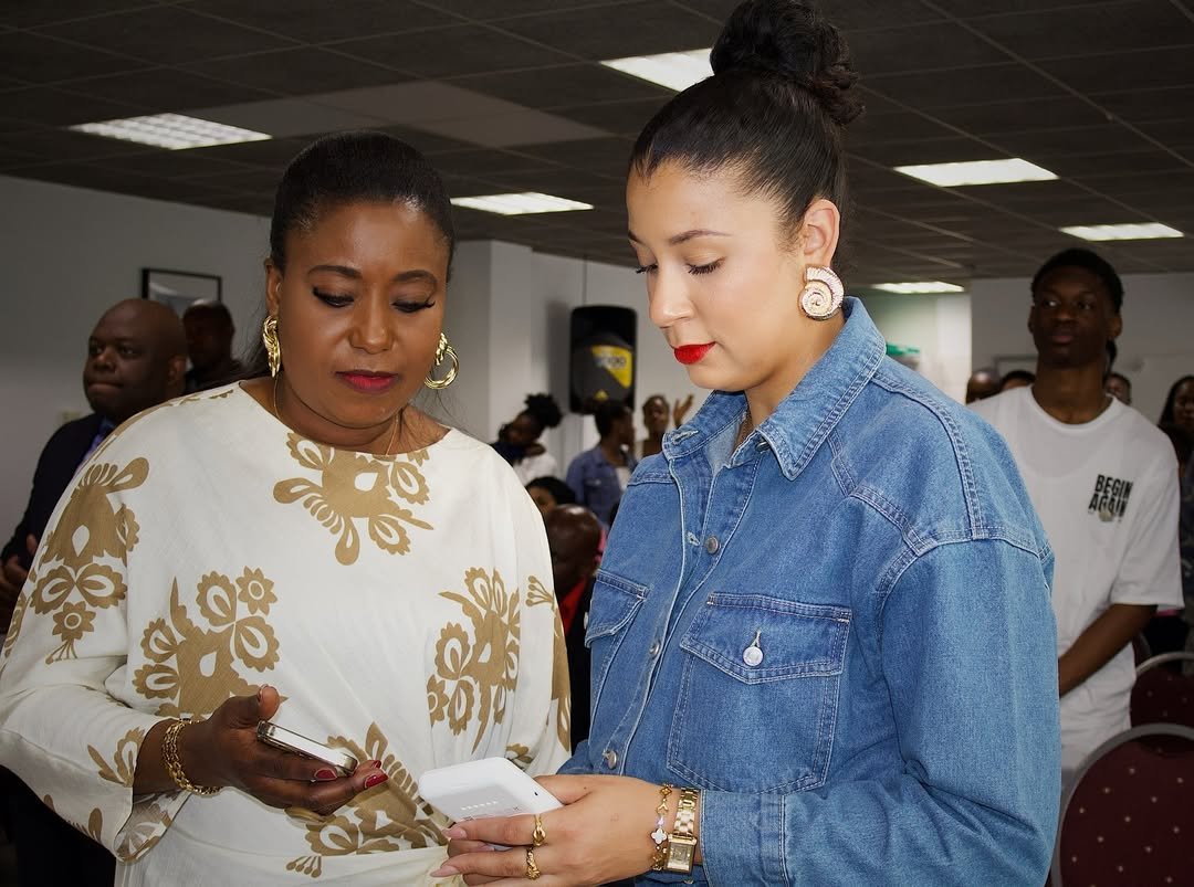 Two women, one in a white and gold floral blouse and the other in a denim jacket, engaged in a conversation while looking at a mobile phone in a busy room.